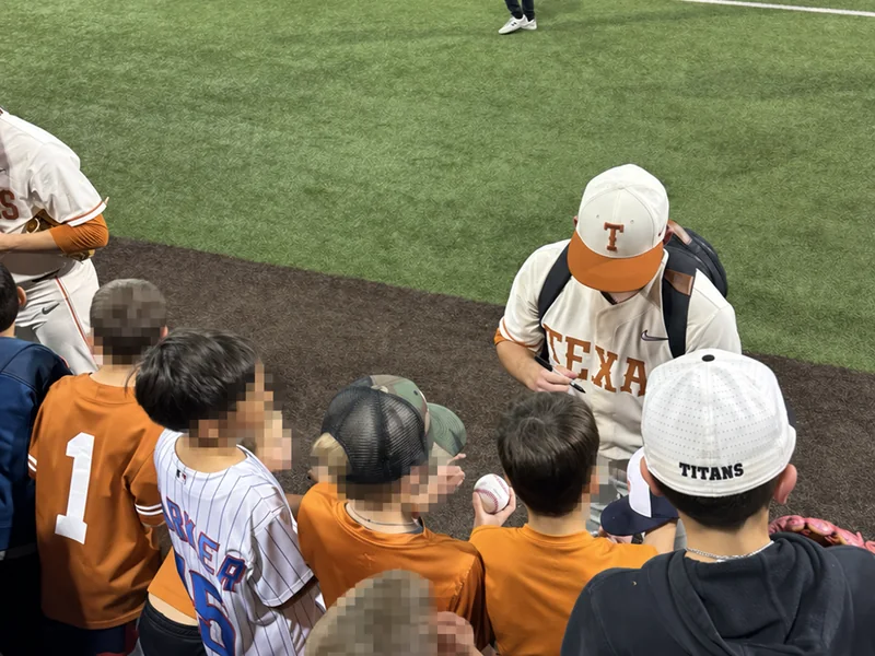 Children gathered along the right-field line while a Texas baseball player signs a ball; children’s faces are obscured for privacy